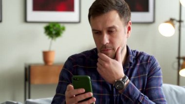 Düşünceli bir insan elinde akıllı bir telefonla kanepede oturur, telefona bakar ve düşünür..