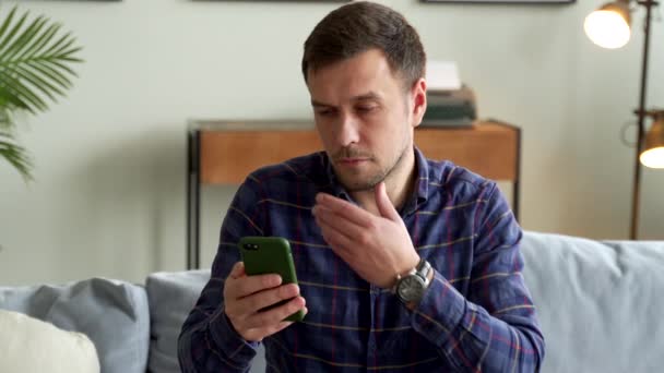 Задумчивый человек сидит на диване со смартфоном в руке, смотрит на телефон и думает:. — стоковое видео