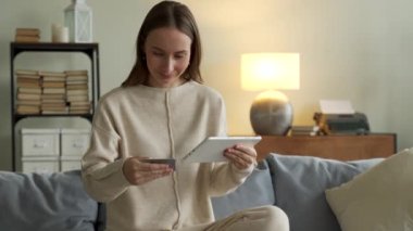 Oturma odasında online alışveriş yapmak için tablet kullanan gülümseyen bir kadın.