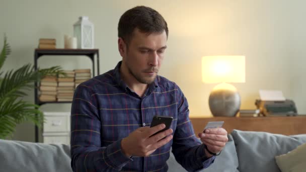 Hombre guapo sentado en el sofá en casa en la sala de estar con teléfono inteligente y tarjeta bancaria de crédito. Pago en línea en concepto de tienda de Internet — Vídeos de Stock