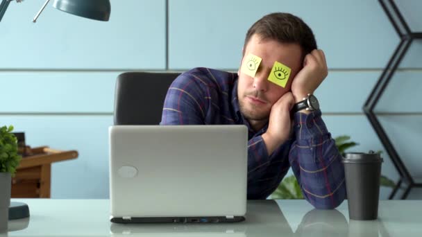 Tired comic male manager pretends working sleeping with stickers on face sits at desk with laptop. – Stock-video