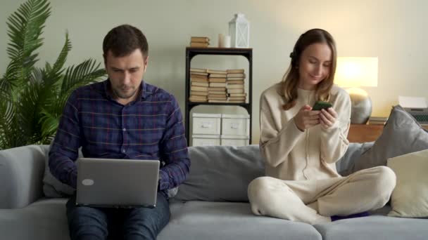 Boldog pár modern kütyükkel otthon. Egy férfi dolgozik egy laptopon, és egy lány fejhallgatóval zenét hallgat egy mobiltelefonon, a kanapén pihen. — Stock videók
