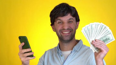 Sarı arka plan stüdyosunda izole edilmiş şaşırmış bir adam. Cep telefonu kullanılıyor. Nakit para hayranı. Dolar banknotları. 