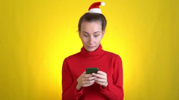 Şaşırmış kadın Noel Baba şapkası cep telefonu kullanıyor sarı arka plan stüdyosunda izole edilmiş mesajlar yazıyor Mutlu yıllar yeni yıl kutlamaları konsepti — Stok video