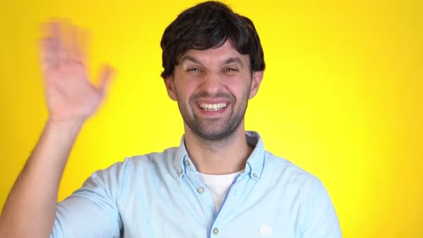 Smiling man isolated on yellow background in studio. Looking camera waving and greeting with hand as notices someone — Stock Video