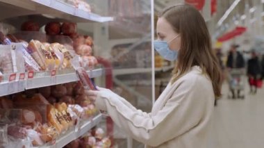 Tıp maskeli genç bir kadın süpermarkette sosis ürünleri seçiyor.
