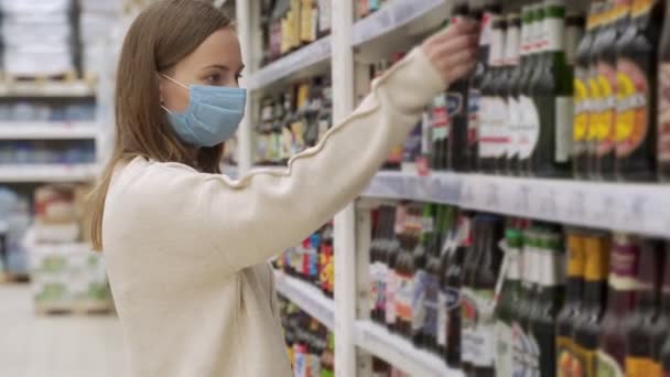 戴着医疗面具的女性从超市货架上拿酒精饮料。在商店里买酒。3.女孩在超市里选择了一种饮料. — 图库视频影像
