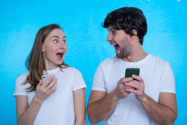 Gens excités positifs homme et femme criant et se regardant tout en utilisant les deux téléphones mobiles isolés sur fond bleu — Photo
