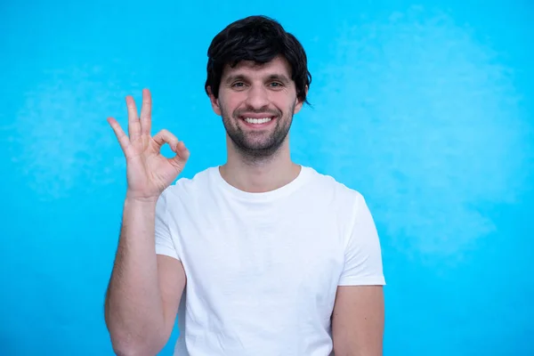 Hombre sonriente mostrando signo de ok y mirando a la cámara sobre fondo azul —  Fotos de Stock