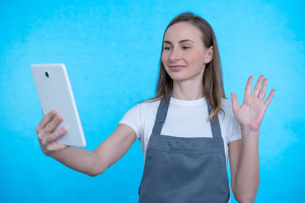 Una mujer sonríe y agita una webcam tableta durante una videollamada sobre un fondo azul — Foto de Stock