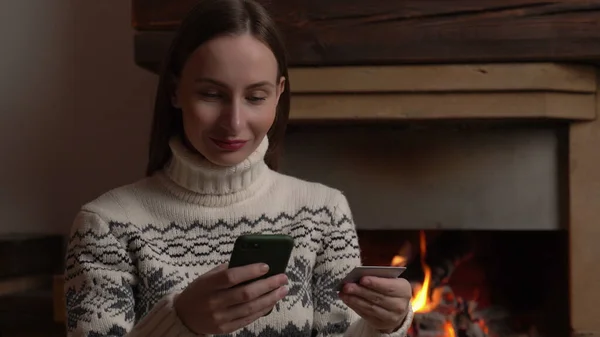女人坐在壁炉边，用智能手机在网上购物 — 图库照片