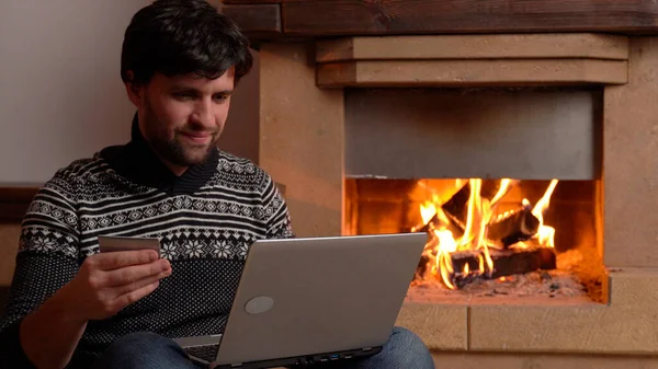 Hombre de compras en línea utilizando el ordenador portátil en casa sentado junto a la chimenea — Foto de Stock