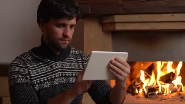 Un hombre emocionado sostiene una tableta y celebra la noticia mientras está sentado junto a la chimenea — Vídeo de stock