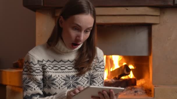 An excited woman holds a tablet and celebrates the news while sitting by the fireplace — Stock Video