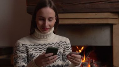 İnternetten akıllı telefon kullanan bir kadın şöminenin yanında oturuyor.