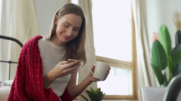 女性はオンラインチャット、オンライン通信のための携帯電話を使用し、テキストメッセージを受信し、毛布で覆われたベッドの上に座ってマグカップを保持します — ストック写真