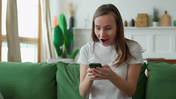 快乐的年轻女士坐在沙发上，用智能手机。兴奋的漂亮女人看着屏幕，在电话游戏中获胜或阅读好消息 — 图库照片