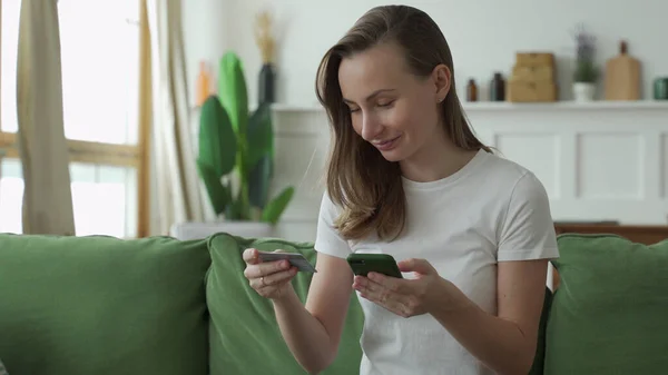 Jonge vrouw die online winkelt met creditcard en smartphone terwijl ze thuis op de bank zit. — Stockfoto