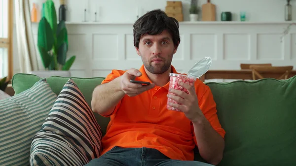 Man sitting on sofa, eating popcorn, holding remote control and switching channels