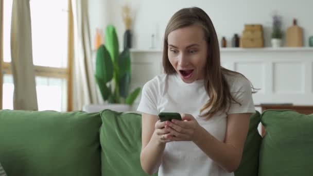 Jovencita alegre sentada en el sofá, usando un smartphone. Emocionado hermosa mujer mira a la pantalla, ganar en el juego de teléfono o leer buenas noticias — Vídeos de Stock