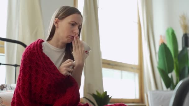 Sjuk kvinna droppar i näsan sitter på sängen och täckt med en röd filt — Stockvideo
