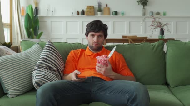 Człowiek siedzący na kanapie, jedzący popcorn, trzymający pilota i przełączający kanały — Wideo stockowe