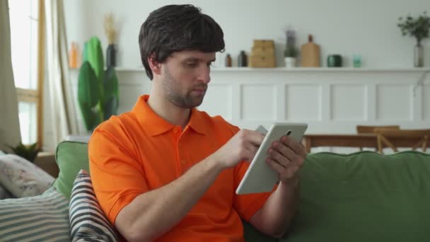 Las manos de los hombres sostienen una tableta, un hombre se sienta en una habitación con una tableta pasa su tiempo libre charlando en línea, sitios web de navegación — Vídeo de stock