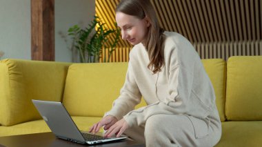 Dizüstü bilgisayarda çalışan, evdeki sarı koltukta oturan bir kadın.