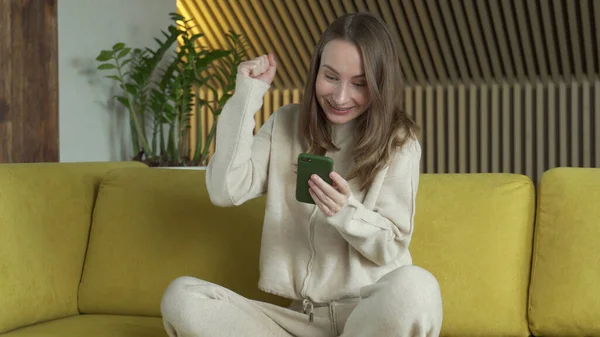 Feliz mujer ganadora sosteniendo el mensaje de lectura de teléfonos inteligentes celebrando la victoria móvil, utilizando el teléfono celular sentarse en el sofá en casa. — Foto de Stock