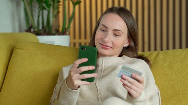 Mujer comprando en línea con una tarjeta de crédito y un teléfono inteligente sentado en un sofá amarillo en casa — Foto de Stock