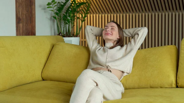 Mujer joven con las manos detrás de la cabeza relajándose en el acogedor sofá amarillo en casa, chica inclinada hacia atrás, estirándose en un cómodo sofá en la sala de estar — Foto de Stock