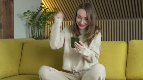 Felice donna vincitrice tenendo smartphone messaggio di lettura che celebra la vittoria mobile, utilizzando il cellulare sedersi sul divano a casa. — Video Stock