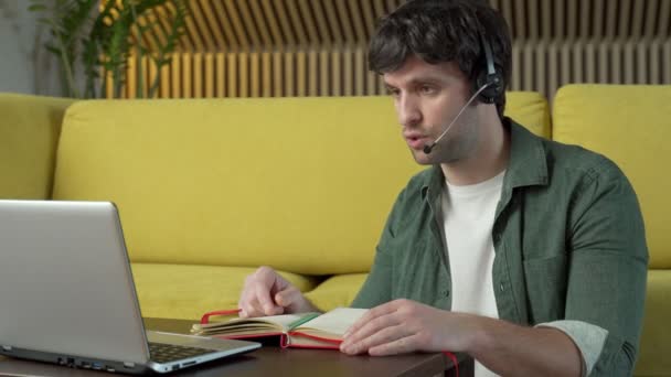 Mladý muž ve sluchátkách sedí doma na žluté pohovce a mluví na videu na notebooku — Stock video