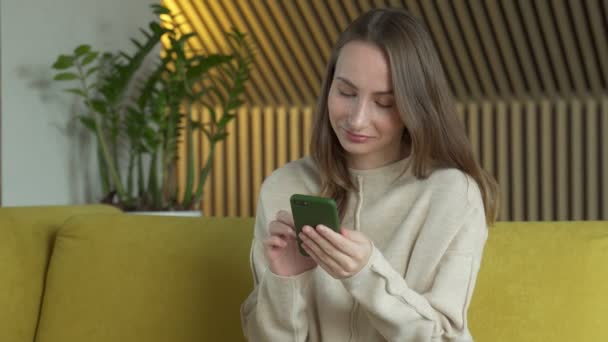 Donna utilizza uno smartphone mentre seduto su un divano giallo nel suo soggiorno. Una donna usa uno smartphone per navigare in Internet a casa. — Video Stock