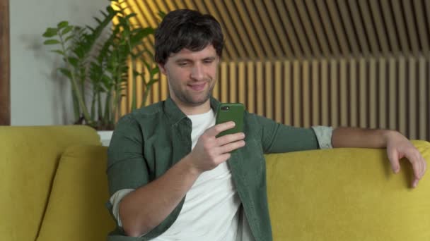 Młody mężczyzna w zielonej koszuli używa smartfona w domu na żółtej kanapie — Wideo stockowe