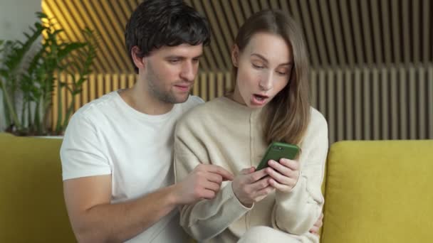 Manželský pár při pohledu na obrazovce smartphone, slaví on-line loterie vyhrát oznámení. Mladí manželé pocit nadšení z sms s dobrou zprávou, sedí na pohovce. — Stock video