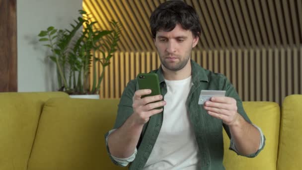 Muž držící smartphone a kreditní karty nakupování on-line nákup v internetovém obchodě sedí na žluté pohovce doma. — Stock video