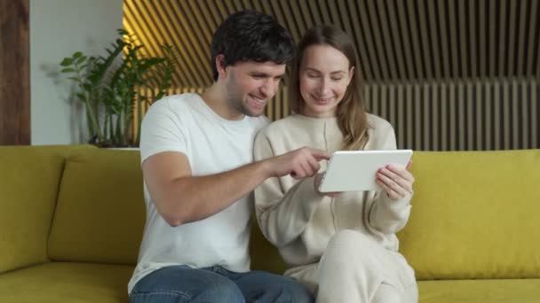 Jovem casal assistindo conteúdo de mídia online em um tablet sentado em um sofá em casa — Vídeo de Stock
