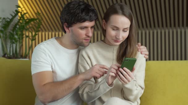 Супружеская пара смотрит на экран смартфона, празднует победу в онлайн-лотерее уведомления. Молодые супруги взволнованы по поводу смс с хорошими новостями, сидя на диване. — стоковое видео