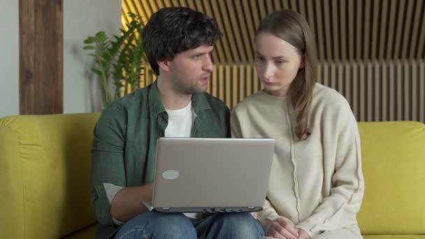 年轻夫妇坐在舒适的黄色沙发上，看着笔记本电脑的屏幕，胜利地在网上赢了彩票，彼此拥抱 — 图库视频影像