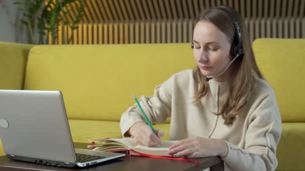 Kulaklık takmış bir kadın evdeki sarı koltukta oturuyor ve dizüstü bilgisayarla görüntülü sohbet ediyor.. — Stok video