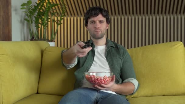 Mężczyzna przed telewizorem oglądający mecz sportowy lub film, siedzący na żółtej kanapie i jedzący popcorn — Wideo stockowe