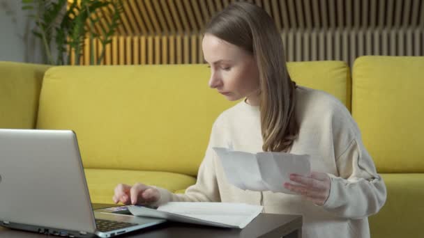 Mladá žena sedí na podlaze vedle žluté pohovky u stolu s papírovými účtenkami a cítí se vystresovaná ze splátek bankovních půjček — Stock video