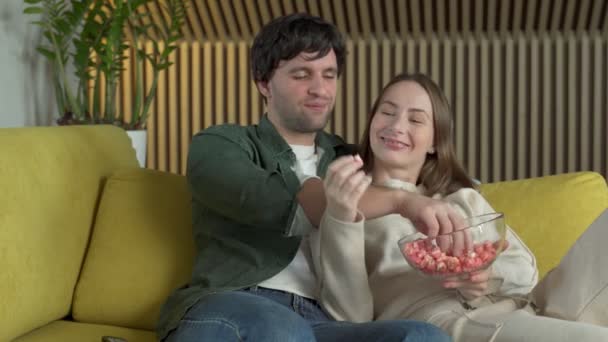 Jeune couple embrassant assis sur un canapé jaune regarder la comédie ayant collation avec une assiette de maïs soufflé — Video