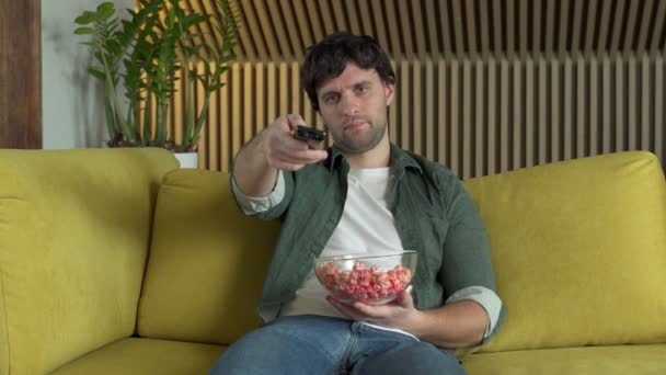 A man in front of the TV watching a sports game or a movie, sitting on a yellow sofa and eating popcorn — Stock Video