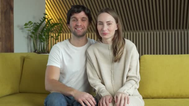 Портрет жены и мужа, размахивающих руками, смотрящих в камеру и говорящих "привет", сидящих на диване в гостиной — стоковое видео