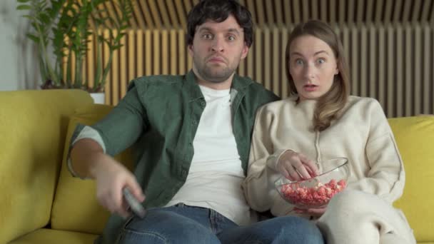 Young couple is sitting on a yellow sofa eating popcorn and watching a movie with a shocked expression on their faces — Stock Video