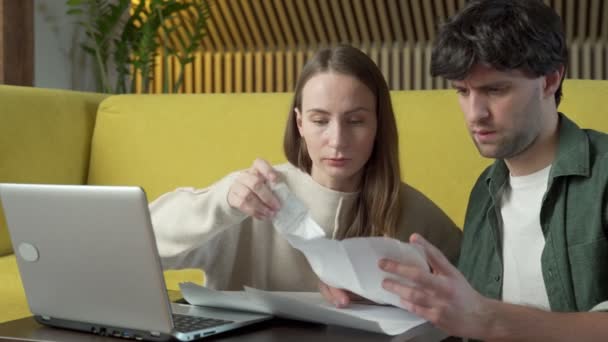 Молодая пара сидит дома на жёлтом диване и управляет бюджетом с помощью ноутбука — стоковое видео