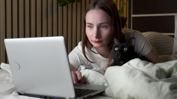 Donna attraente con un gatto nero utilizza un computer portatile mentre sdraiato sul letto — Video Stock