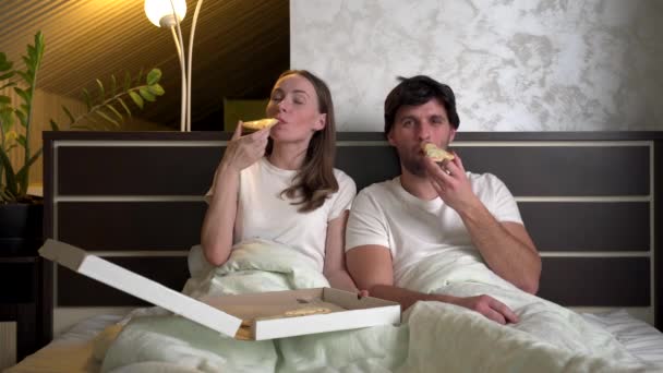 Young couple watches TV in bed and eats pizza in the evening — Stock Video